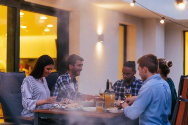 Bir grup genç insan akşamları modern bir evin terasında yemek yiyorlar. Dostlar ve aile için eğlence. Tatil kutlamaları, barbekülü düğünler..