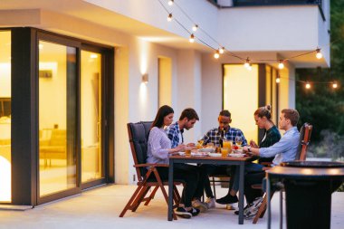 Bir grup genç insan akşamları modern bir evin terasında yemek yiyorlar. Dostlar ve aile için eğlence. Tatil kutlamaları, barbekülü düğünler..