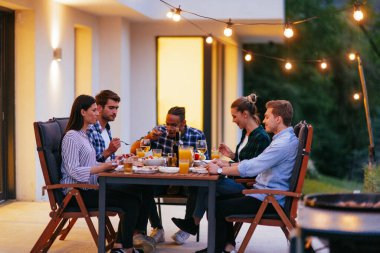 Bir grup genç insan akşamları modern bir evin terasında yemek yiyorlar. Dostlar ve aile için eğlence. Tatil kutlamaları, barbekülü düğünler..