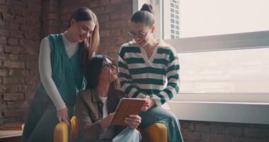 Kadın iş adamları pencere kenarında iş birliği yapıyor, fikirleri tartışıyor ve tablet üzerinde beyin fırtınası yapıyorlar. Modern bir ofiste işbirliği, yaratıcılık ve takım çalışması kavramı