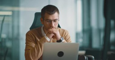 Akıllı görünümlü odaklanmış iş adamı bir dizüstü bilgisayarda çalışıyor ve düşünüyor. Milenyum dönemi aile bütçesini yönetmek, finansal evrak işlerini halletmek ve modern bir açık uzay ofisindeki dizüstü bilgisayardaki harcamaları takip etmek.