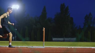 Güçlü Erkek Sporcu bir engele doğru koşuyor, bir yarışta koşarken bariyerin üzerinden yüksek hızda atlıyor. Hız Rampası Efektli Süper Yavaş Sinema Görüntüsü.
