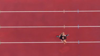 İşaretli bir yarış pisti boyunca koşan, pistin sınırlarını gösteren, hız, odaklanma ve hareket halindeki hassasiyeti vurgulayan tek kişilik bir erkek atletin havadan üst görüntüsü. Sinematik İHA yavaş çekim