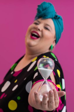 Kendine güvenen bir kadının portresi, eski bir başörtüsü ve renkli kıyafetiyle, zamanın ve odak noktasının sembolü olarak yüzüne bir kum saati tutuyor, parlak pembe bir arka planı var.. 