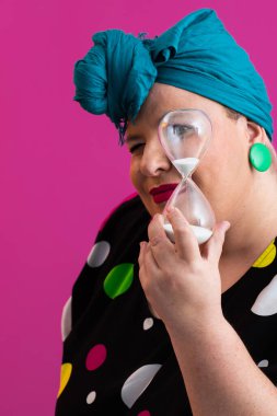 Kendine güvenen bir kadının portresi, eski bir başörtüsü ve renkli kıyafetiyle, zamanın ve odak noktasının sembolü olarak yüzüne bir kum saati tutuyor, parlak pembe bir arka planı var.. 