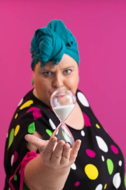 Kendine güvenen bir kadının portresi, eski bir başörtüsü ve renkli kıyafetiyle, zamanın ve odak noktasının sembolü olarak yüzüne bir kum saati tutuyor, parlak pembe bir arka planı var.. 