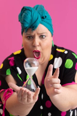 Kendine güvenen bir kadının portresi, eski bir başörtüsü ve renkli kıyafetiyle, zamanın ve odak noktasının sembolü olarak yüzüne bir kum saati tutuyor, parlak pembe bir arka planı var.. 