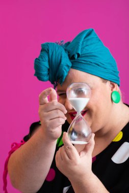 Kendine güvenen bir kadının portresi, eski bir başörtüsü ve renkli kıyafetiyle, zamanın ve odak noktasının sembolü olarak yüzüne bir kum saati tutuyor, parlak pembe bir arka planı var.. 