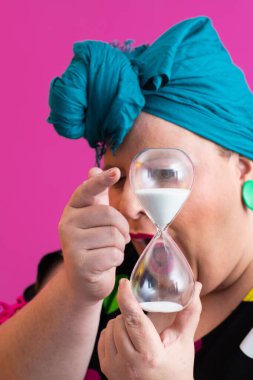 Kendine güvenen bir kadının portresi, eski bir başörtüsü ve renkli kıyafetiyle, zamanın ve odak noktasının sembolü olarak yüzüne bir kum saati tutuyor, parlak pembe bir arka planı var.. 