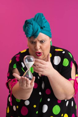 Kendine güvenen bir kadının portresi, eski bir başörtüsü ve renkli kıyafetiyle, zamanın ve odak noktasının sembolü olarak yüzüne bir kum saati tutuyor, parlak pembe bir arka planı var.. 