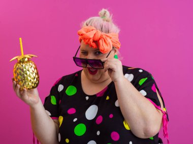 Kendine güveni tam ve eskimiş bir kadın elinde altın bir ananas bardağı tutarken parlak pembe bir arka plana karşı tavır takınıyor.. 