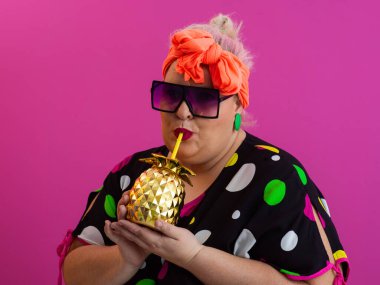 Kendine güveni tam ve eskimiş bir kadın elinde altın bir ananas bardağı tutarken parlak pembe bir arka plana karşı tavır takınıyor.. 