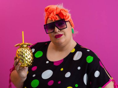 Kendine güveni tam ve eskimiş bir kadın elinde altın bir ananas bardağı tutarken parlak pembe bir arka plana karşı tavır takınıyor.. 