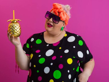 Kendine güveni tam ve eskimiş bir kadın elinde altın bir ananas bardağı tutarken parlak pembe bir arka plana karşı tavır takınıyor.. 