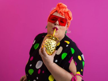 Kendine güveni tam ve eskimiş bir kadın elinde altın bir ananas bardağı tutarken parlak pembe bir arka plana karşı tavır takınıyor.. 