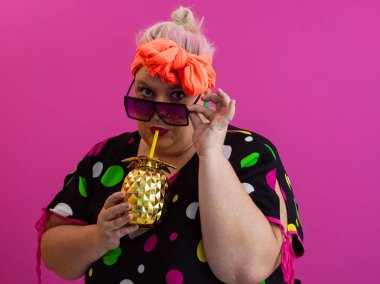 Kendine güveni tam ve eskimiş bir kadın elinde altın bir ananas bardağı tutarken parlak pembe bir arka plana karşı tavır takınıyor.. 