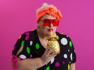 Kendine güveni tam ve eskimiş bir kadın elinde altın bir ananas bardağı tutarken parlak pembe bir arka plana karşı tavır takınıyor.. 