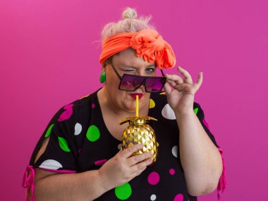 Kendine güveni tam ve eskimiş bir kadın elinde altın bir ananas bardağı tutarken parlak pembe bir arka plana karşı tavır takınıyor.. 