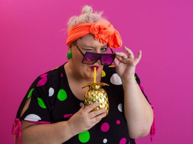 Kendine güveni tam ve eskimiş bir kadın elinde altın bir ananas bardağı tutarken parlak pembe bir arka plana karşı tavır takınıyor.. 