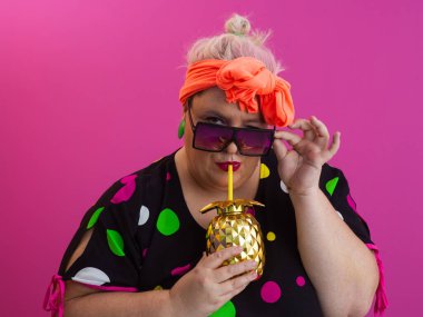 Kendine güveni tam ve eskimiş bir kadın elinde altın bir ananas bardağı tutarken parlak pembe bir arka plana karşı tavır takınıyor.. 