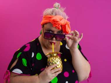 Kendine güveni tam ve eskimiş bir kadın elinde altın bir ananas bardağı tutarken parlak pembe bir arka plana karşı tavır takınıyor.. 