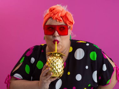 Kendine güveni tam ve eskimiş bir kadın elinde altın bir ananas bardağı tutarken parlak pembe bir arka plana karşı tavır takınıyor.. 