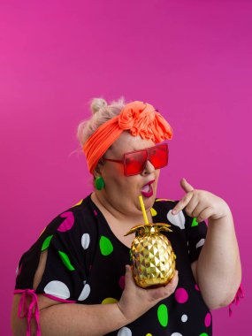 Kendine güveni tam ve eskimiş bir kadın elinde altın bir ananas bardağı tutarken parlak pembe bir arka plana karşı tavır takınıyor.. 