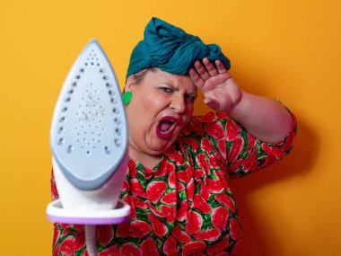 Artı canlı karpuz desenli elbisesi ve deniz mavisi başlıklı, elinde elektrikli ütüyle parlak turuncu arka planda komik bir ifadeyle bağıran iri yarı bir kadın. Kavramsal ve renkli