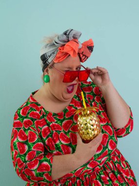 Karpuz desenli bir elbise giymiş tuhaf bir kadın ve elinde altın ananas şeklindeki bir kupayla kırmızı güneş gözlüklerinin üzerinden neşeyle bakan bir kupa tutuyor.. 