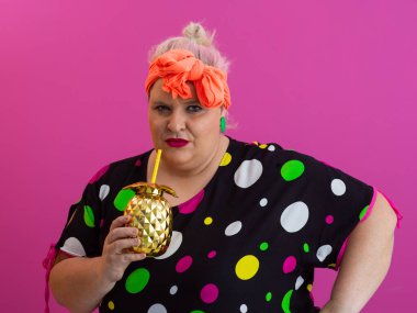 Kendine güveni tam ve eskimiş bir kadın elinde altın bir ananas bardağı tutarken parlak pembe bir arka plana karşı tavır takınıyor.. 