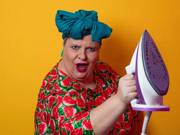 Artı canlı karpuz desenli elbisesi ve deniz mavisi başlıklı, elinde elektrikli ütüyle parlak turuncu arka planda komik bir ifadeyle bağıran iri yarı bir kadın. Kavramsal ve renkli