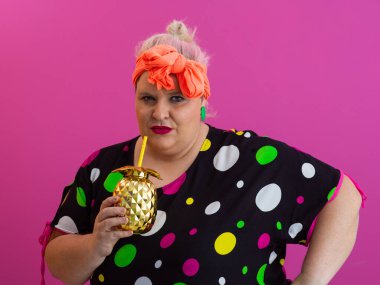 Kendine güveni tam ve eskimiş bir kadın elinde altın bir ananas bardağı tutarken parlak pembe bir arka plana karşı tavır takınıyor.. 
