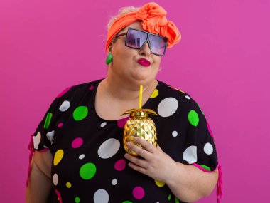 Kendine güveni tam ve eskimiş bir kadın elinde altın bir ananas bardağı tutarken parlak pembe bir arka plana karşı tavır takınıyor.. 