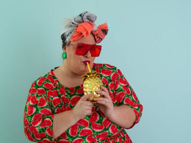 Karpuz desenli bir elbise giymiş tuhaf bir kadın ve elinde altın ananas şeklindeki bir kupayla kırmızı güneş gözlüklerinin üzerinden neşeyle bakan bir kupa tutuyor.. 