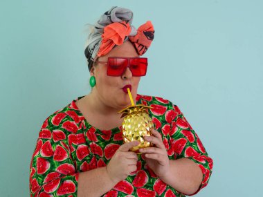 Karpuz desenli bir elbise giymiş tuhaf bir kadın ve elinde altın ananas şeklindeki bir kupayla kırmızı güneş gözlüklerinin üzerinden neşeyle bakan bir kupa tutuyor.. 