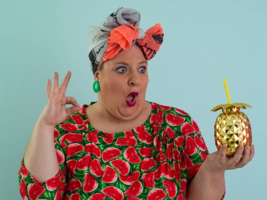 Karpuz desenli bir elbise giymiş tuhaf bir kadın ve elinde altın ananas şeklindeki bir kupayla kırmızı güneş gözlüklerinin üzerinden neşeyle bakan bir kupa tutuyor.. 