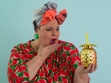 Karpuz desenli bir elbise giymiş tuhaf bir kadın ve elinde altın ananas şeklindeki bir kupayla kırmızı güneş gözlüklerinin üzerinden neşeyle bakan bir kupa tutuyor.. 