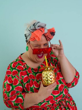 Karpuz desenli bir elbise giymiş tuhaf bir kadın ve elinde altın ananas şeklindeki bir kupayla kırmızı güneş gözlüklerinin üzerinden neşeyle bakan bir kupa tutuyor.. 