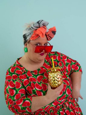 Karpuz desenli bir elbise giymiş tuhaf bir kadın ve elinde altın ananas şeklindeki bir kupayla kırmızı güneş gözlüklerinin üzerinden neşeyle bakan bir kupa tutuyor.. 