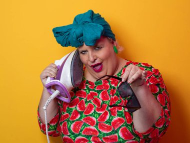 Artı canlı karpuz desenli elbisesi ve deniz mavisi başlıklı, elinde elektrikli ütüyle parlak turuncu arka planda komik bir ifadeyle bağıran iri yarı bir kadın. Kavramsal ve renkli