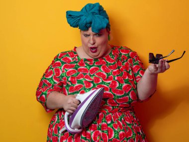 Artı canlı karpuz desenli elbisesi ve deniz mavisi başlıklı, elinde elektrikli ütüyle parlak turuncu arka planda komik bir ifadeyle bağıran iri yarı bir kadın. Kavramsal ve renkli