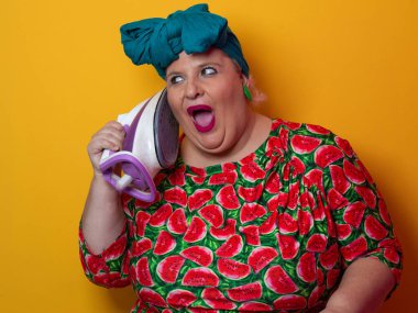 Artı canlı karpuz desenli elbisesi ve deniz mavisi başlıklı, elinde elektrikli ütüyle parlak turuncu arka planda komik bir ifadeyle bağıran iri yarı bir kadın. Kavramsal ve renkli