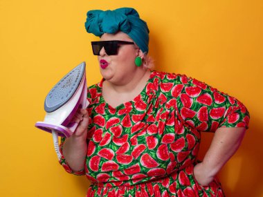 Artı canlı karpuz desenli elbisesi ve deniz mavisi başlıklı, elinde elektrikli ütüyle parlak turuncu arka planda komik bir ifadeyle bağıran iri yarı bir kadın. Kavramsal ve renkli