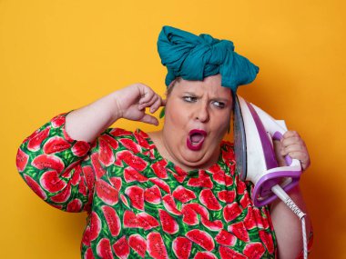 Artı canlı karpuz desenli elbisesi ve deniz mavisi başlıklı, elinde elektrikli ütüyle parlak turuncu arka planda komik bir ifadeyle bağıran iri yarı bir kadın. Kavramsal ve renkli