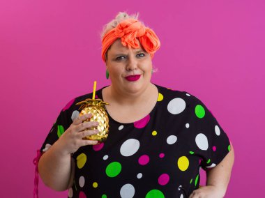 Kendine güveni tam ve eskimiş bir kadın elinde altın bir ananas bardağı tutarken parlak pembe bir arka plana karşı tavır takınıyor.. 