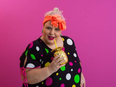 Kendine güveni tam ve eskimiş bir kadın elinde altın bir ananas bardağı tutarken parlak pembe bir arka plana karşı tavır takınıyor.. 