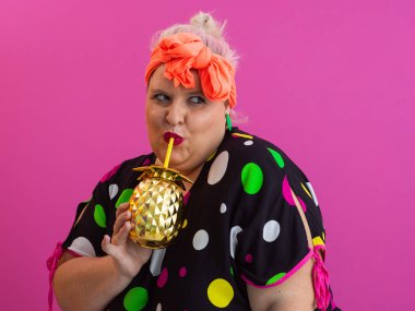 Kendine güveni tam ve eskimiş bir kadın elinde altın bir ananas bardağı tutarken parlak pembe bir arka plana karşı tavır takınıyor.. 