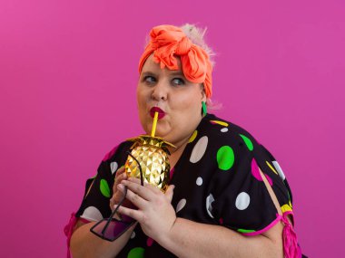 Kendine güveni tam ve eskimiş bir kadın elinde altın bir ananas bardağı tutarken parlak pembe bir arka plana karşı tavır takınıyor.. 