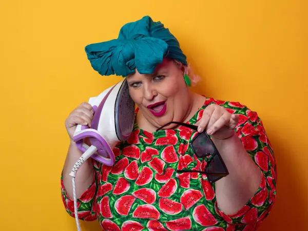 Artı canlı karpuz desenli elbisesi ve deniz mavisi başlıklı, elinde elektrikli ütüyle parlak turuncu arka planda komik bir ifadeyle bağıran iri yarı bir kadın. Kavramsal ve renkli