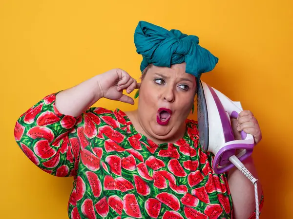 Artı canlı karpuz desenli elbisesi ve deniz mavisi başlıklı, elinde elektrikli ütüyle parlak turuncu arka planda komik bir ifadeyle bağıran iri yarı bir kadın. Kavramsal ve renkli